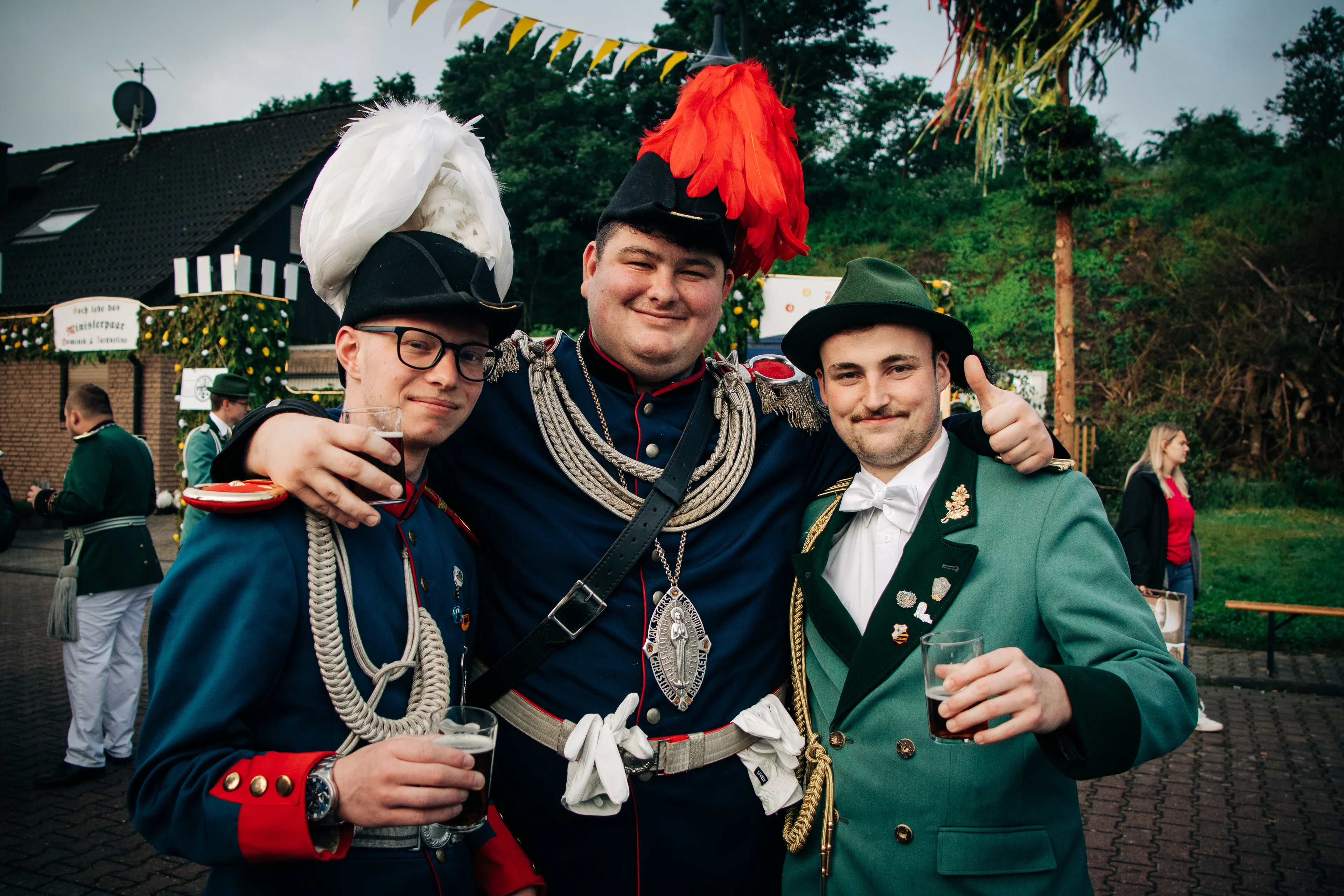 Drei lächelnde Schützenbrüder in historischen Uniformen, teilweise mit prächtigen Federhüten und Königssilber, stehen gesellig zusammen und halten Gläser mit Altbier.