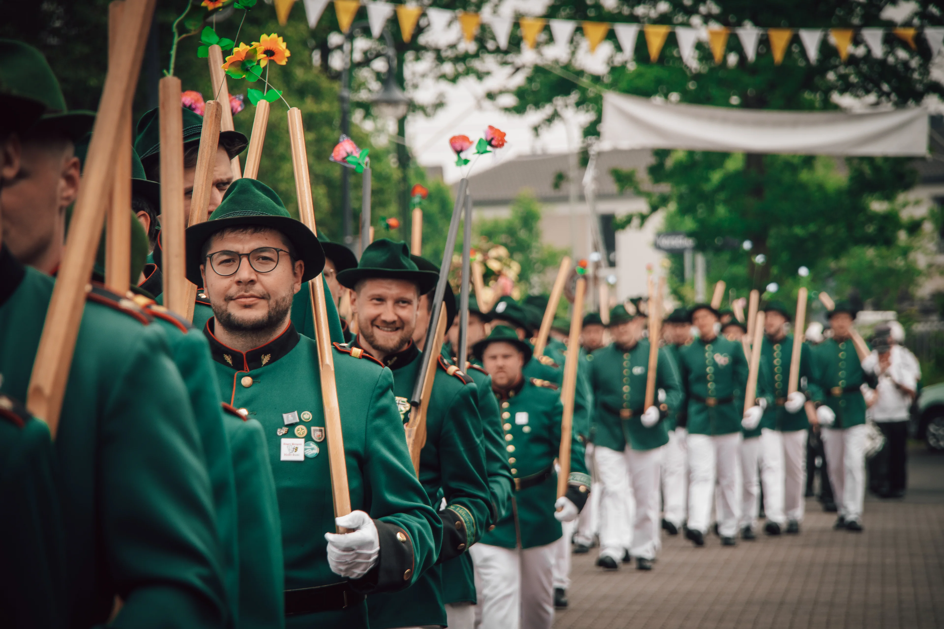 Schützen der St. Katharina Junggesellen Bruderschaft marschieren in grünen Uniformen und mit blumengeschmückten Holzgewehren bei einer Parade.