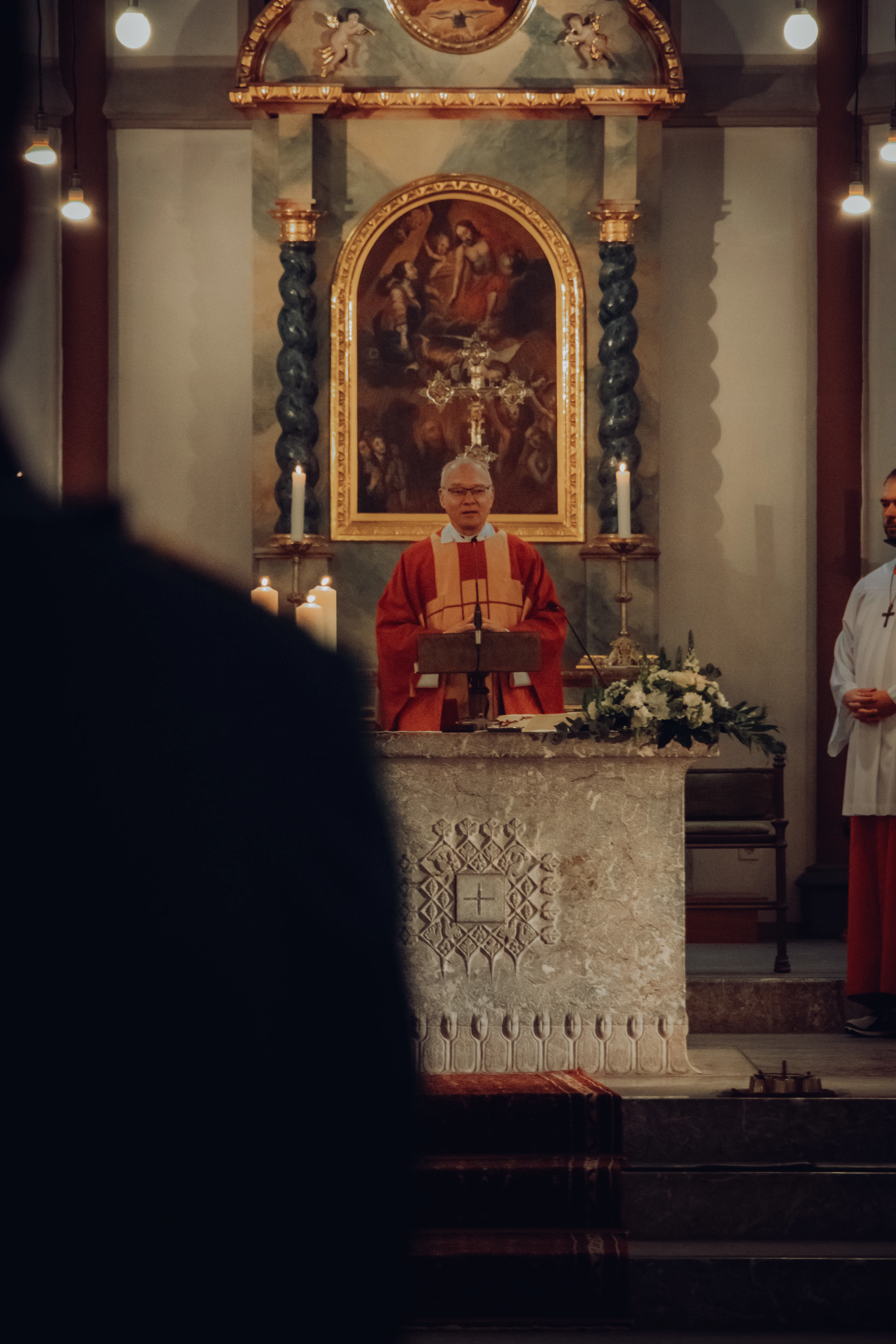 Ein Priester in rotem Messgewand hält eine Predigt am Altar einer festlich beleuchteten Kirche.